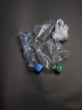 Koyu gri bir yüzeye dağılmış çeşitli ezilmiş plastik şişe ve bardakların yukarıdan aşağı görüntüsü. Görüntü, atılan tek kullanımlık eşyalar sonrasında ortaya çıkan plastik atıkların çevresel etkilerini vurguluyor. Kompozisyon dokuları vurguluyor