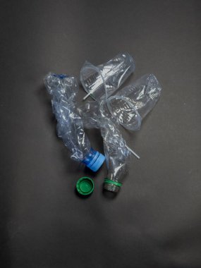 Koyu gri bir yüzeye saçılmış buruşmuş plastik şişeler ve bardaklar. Yeşil ve mavi şişe kapakları da görülebilir. Görüntü, plastik atık, çevre kirliliği ve geri dönüşüm ve atık idarecilerine duyulan ihtiyacı vurguluyor