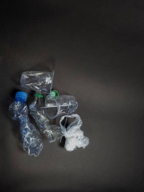 Tepeden çekilen resimde, koyu gri bir arka plana saçılmış şişe, bardak ve plastik torba da dahil olmak üzere çeşitli plastik atık parçaları görülüyor. Kompozisyon, plastik kirliliğinin yaygınlığını ve geri dönüşüm ve çevreye duyulan ihtiyacı vurguluyor