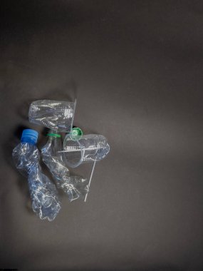 Yukarıdan aşağıya atılan bir çekim, koyu gri arka planda şişeler, bardaklar ve kapaklar da dahil olmak üzere atılmış plastik atıkları yakalar. Plastiğin buruşuk ve kullanılmış görünümü, çevre kirliliği ve geri dönüşüm veya atık azaltma çabalarına duyulan ihtiyacı ortaya koyuyor. Th
