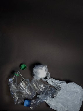 Tepetaklak bir karede şişeler, bardaklar ve koyu bir yüzeye saçılmış bir poşet gibi çeşitli plastik atık eşyalar sergileniyor. Bu görüntü kirlilik ve çevre sorunlarının görsel bir temsilidir ve geri dönüşüm ve atık azaltma ihtiyacını vurgular