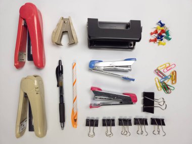 Bu resim, düz beyaz bir arkaplanda düzgün bir şekilde düzenlenmiş temel ofis malzemelerinin bir koleksiyonunu gösteriyor. Kompozisyonda zımba, zımba çıkarıcı, delgeç, raptiye, ataç, ataç, kalem ve çizgili kalem yer alıyor. Temiz giriş.