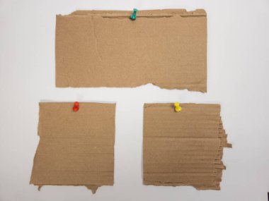 Üç parça yırtık karton renkli raptiyelerle beyaz bir yüzeye tutturulmuş. Düzenleme basit ve temiz bir estetik yaratıyor. Görüntü, kartondaki dokuyu ve doğal tonlar ile parlak rozet eş arasındaki kontrastı gösteriyor.