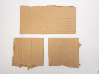 Bu üstten çekimde beyaz arka planda yırtık kenarlı üç parça kahverengi karton görünüyor. Karton parçalar farklı boyutlarda ve şekillerde. Kenarları da düzgün değil. Bu da yırtıldığını ya da kesildiğini gösteriyor. Görüntü iyi aydınlatılmış.