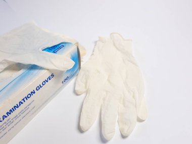 Yakın plan çekimde bir kutu sınav eldiveni ve yanında tek eldiven var. Hepsi beyaz arka planda. Eldivenler açık renkli bir malzemeden yapılmış ve kutu eldivenlerle ilgili detaylarla etiketlenmiş. Bu görüntü tıbbı temsil ediyor.