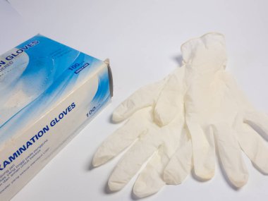 Beyaz bir yüzey üzerinde bir kutu tıbbi inceleme eldiveni ve bir çift eldiven bulunan bir stüdyo resmi. Eldivenler soluk sarı bir maddeden yapılmış ve kutuda mavi ve beyaz markalar var. Görüntü iyi aydınlatılmış ve minimalist bir aesth 'e sahip.