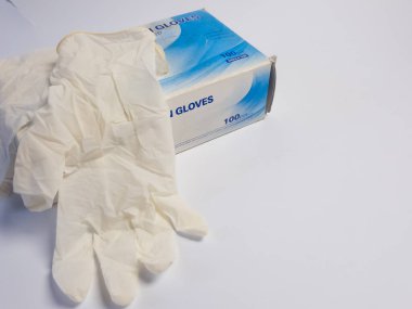 Bir çift tıbbi eldiven, beyaz arka planda izole edilmiş bir eldiven kutusunun yanında yatıyor. Eldivenler açık renkli bir maddeden yapılmış ve kullanılmamış gibi görünüyor. Kutu metin ve grafiklerle etiketlenmiş. Kompozisyon basit ve gl 'ye odaklı.