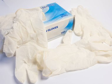 Bu resim, beyaz bir yüzeye dağılmış birkaç eldivenli tek kullanımlık tıbbi eldiven kutusunu gösteriyor. Eldivenler şeffaf bir malzemeden yapılmıştır. Kompozisyon yakın çekim, eldivenlerin ve kutunun detaylarını vurguluyor.. 