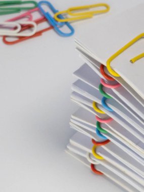 Yakın plan çekimde renkli ataçlarla tutturulmuş beyaz kağıtlar sergileniyor. Belgelerin kenarlarına çeşitli ataç renkleri iliştirilmiştir. Kompozisyon temiz ve basit, düz beyaz bir arkaplan üzerine kurulmuş. Resim eşleşmesi