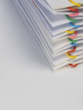 Yakın plan çekimde, renkli ataçlarla düzgünce bir araya getirilmiş bir yığın beyaz kağıt dökümanı yer alıyor. Kompozisyon, belgelerin organize düzenlenmesini, açık beyaz kağıdı ve canlı ataçları vurguluyor. Resim eşleşmesi