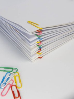 Beyaz arka planda renkli ataçlarla tutturulmuş bir yığın kağıt. Çeşitli renkli ataçlar alta saçılırken, diğerleri kağıt yığınının içindeki bölümleri ayırmak için kullanılıyor. Görüntü iyi aydınlatılmış ve temiz., 