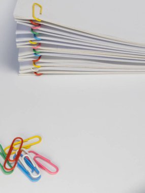 Beyaz bir yüzeye yerleştirilmiş renkli ataçlarla tutturulmuş beyaz kağıt yığınının yakın plan çekimi. Görüntü, çıtır beyaz kağıt ile ataçların canlı renkleri arasındaki zıtlığı vurgular. Kompozisyon basit ve temiz., 