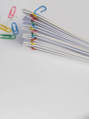 Renkli ataçlarla tutturulmuş beyaz kağıt yığınının yakın plan çekimi. Beyaz arka planda yığının etrafına çeşitli renkli ataçlar saçıldı. Kompozisyon temiz ve minimalist. Organize doğayı vurguluyor.