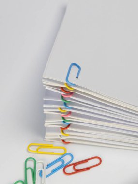 Çeşitli renkli ataçlarla korunan beyaz kağıtların yakın plan çekimi. Birkaç tane gevşek ataç yığının altındaki beyaz yüzeye saçılmış. Görüntü iyi aydınlatılmış, temiz ve basit bir estetik, iş ve ofis için uygun.