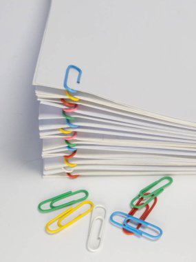 Yakın plan bir stüdyo çekiminde, çeşitli renkli ataçlarla tutturulmuş beyaz kağıtlar ve beyaz yüzeye saçılmış birkaç şarjör bulunur. Görüntü, ofis malzemelerini ve organizasyon kavramlarını vurguluyor, renkleri vurguluyor ve c