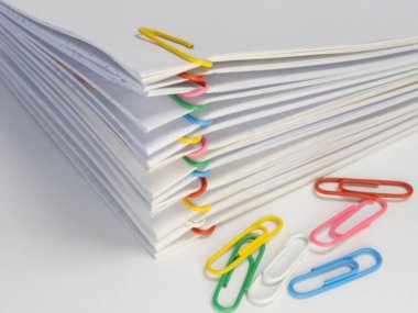 Çeşitli renkli ataçlarla tutturulmuş beyaz kağıt yığınının yakın plan fotoğrafı. Farklı renkte birkaç ataç yığının yanına saçılmış. Görüntü iyi aydınlatılmış, beyaz kağıt arasındaki kontrastı vurguluyor.