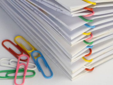 Çeşitli renkli ataçlarla tutturulmuş beyaz kağıt yığınının yakın plan çekimi. Görüntü beyaz bir yüzeyde çekiliyor, temiz ve düzenli bir estetik oluşturuyor. Kağıtlar kırmızı, sarı, yeşil, pembe ve mavi renklidir. Th