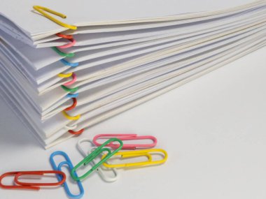 Beyaz arka planda çeşitli renkli ataçları olan beyaz bir kağıt yığını tasvir edilir. Görüntü ofis malzemeleri, organizasyon ve belge yönetimini ön plana çıkarıyor. Kağıt yığınına ve canlı ataçlara odaklanıyoruz.