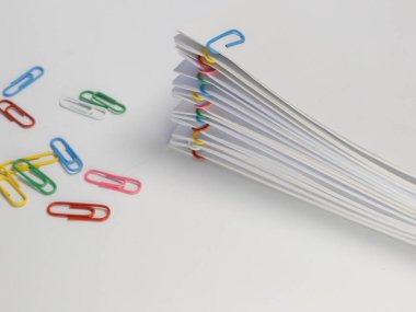 Renkli ataçlarla bir araya getirilmiş beyaz kağıt yığını. Beyaz yüzeye çeşitli renkli ataçlar saçılmıştır. Beyaz kağıt ve inci arasındaki kontrast odaklı, temiz, minimalist bir estetiğe sahiptir