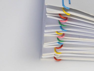 Bu resim renkli ataçlarla bir araya getirilmiş beyaz kağıt yığınının yakın plan görüntüsünü içeriyor. Kağıt ataçları çeşitli renklerde, beyaz kağıda karşı görsel olarak çekici bir karşıtlık yaratıyor. Kompozisyon temiz ve minimal.