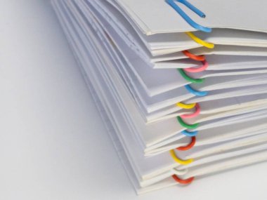 Çeşitli renkli ataçlarla tutturulmuş beyaz kağıt çarşafların yakın plan çekimi. Kağıtlar düzgünce istiflenmiş ve klipler eşit aralıklarla yerleştirilmiş. Bu da görsel olarak çekici bir kompozisyon oluşturuyor. Arka plan temiz, beyaz bir yüzey. Bu...