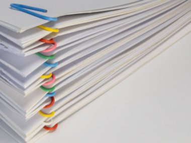 Çeşitli renkli ataçlarla bir arada tutulan bir yığın beyaz kağıt. Kağıtlar düzgünce istiflenmiş. Organizasyon ve belge yönetimi ihtiyacı var. Ataçlar beyaz babaya zıt bir renk katıyor.