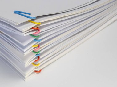 Renkli ataçlarla tutturulmuş beyaz kağıtların yakın plan çekimi. Kağıtlar düzgünce istiflenmiş ve ataçlar da sıraya dizilmiş. Arka plan düz beyaz bir yüzey. Görüntü bir düzen ve doküman duygusu iletir