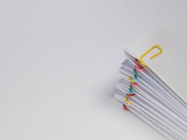 Yakın plan çekimde çeşitli renkli ataçlarla tutturulmuş beyaz kağıt çarşaf yığını yer alır. Kağıtlar, düz beyaz bir arkaplana karşı hafif bir açıyla düzenlenmiş, temiz ve minimalist bir estetik yaratıyor. Kompozisyon yüksekliği