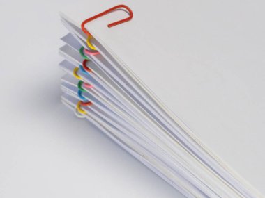 Çeşitli renkli ataçlarla tutturulmuş beyaz kağıt yığınının yakın plan fotoğrafı. Kağıt, tek tek çarşafları gösteren bir şekilde yayılmış. Görüntü düz beyaz bir arkaplanda çekilmiştir. Ataçlar kırmızı, sarı, gr