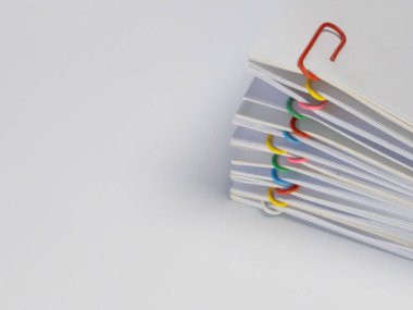 Yakın plan çekimde renkli ataçlarla korunan bir yığın beyaz kağıt sergileniyor. Bu ataçlar kırmızı, sarı, yeşil, pembe ve mavi gibi çeşitli renklerdir ve beyaz kağıtlara karşı görsel bir kontrasttır. Görüntü ya da