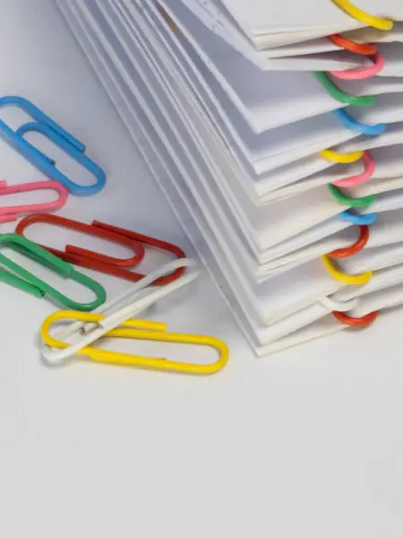 Renkli ataçlarla tutturulmuş beyaz kağıtların yakın plan çekimi. Çeşitli renklerde ataçlar beyaz zemine saçılmış. Bu da organizasyon, evrak ve ofis malzemelerini işaret ediyor. İmajın parlak ve temiz bir ücreti var.