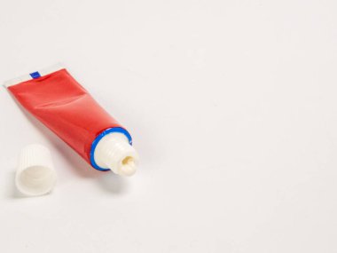 Bu görüntüde beyaz bir yüzey üzerinde duran kapağı açık kırmızı bir krem tüpü bulunur. Tüpü açılı ve küçük bir miktar krem açıdan dışarı çıkıyor. Kompozisyon basit ve temiz, ürüne odaklanmış ve ben...