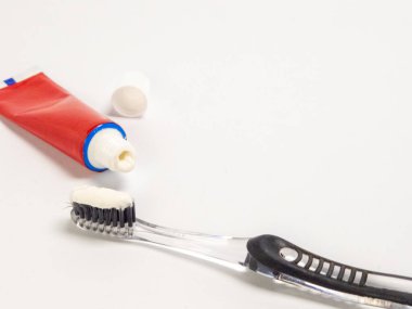 Beyaz arka planda siyah tüyleri ve bir tüp kırmızı diş macunu olan temiz bir diş fırçası bulunmaktadır. Diş macunu fırçaya sıkışıyor ve tüpün kapağı yakınlarda. Görüntü temiz ve parlak, ağız sağlığını temsil ediyor. 