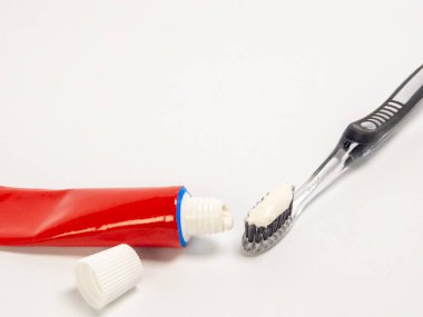 Diş fırçasının diş fırçasının diş fırçasına sürdüğü yakın plan bir fotoğraf, diş macununun kapağı çıkarılmış kırmızı bir tüpün yanında. Eşyalar temiz, beyaz bir arka plana göre ayarlanarak basit ve modern bir estetik oluşturuluyor. Görüntü vurgular 