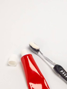 Kapağı açık kırmızı bir diş macunu tüpünün yakın plan çekimi, diş macunlu diş fırçası ve kapağın kendisi. Eşyalar temiz, beyaz bir arkaplanda düzenleniyor. Kompozisyon diş hijyenine ve hazırlıklarına odaklanıyor.
