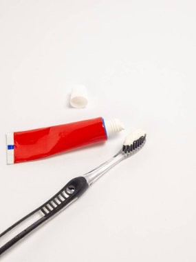 Yakın plan çekimde, üzerinde diş macunu olan bir diş fırçası ve kapağı açık bir kırmızı diş macunu tüpü, hepsi temiz beyaz bir arkaplan üzerine kuruludur. Temiz diş fırçası sapı ve siyah tüyler, beyaz diş macunuyla birlikte görünür. T