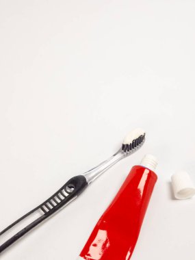 Tüylerinde beyaz diş macunu olan şeffaf siyah bir diş fırçası, kırmızı bir diş macunu tüpü ve beyaz kapağı, hepsi beyaz bir zemine karşı izole edilmiş. Görüntü iyi aydınlatılmış ve temizlik ve hijyen hissi uyandırıyor..