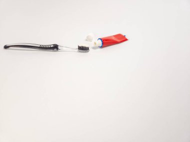 Siyah saplı temiz bir diş fırçası kapaklı kırmızı bir diş macunu tüpünün yanında oturur. Diş fırçasının fırçasında zaten az miktarda diş macunu var. Sahne, basitlik ve temizliği vurgulayan düz beyaz bir arkaplan üzerine kurulmuştur.