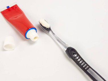 Kapağı açık kırmızı bir diş macunu tüpü ve diş macunlu diş fırçası içeren yakın plan bir çekim, hepsi beyaz arka planda. Bu resim diş bakımını, ağız hijyenini ve diş fırçalamanın günlük rutinini gösteriyor. Şey... 
