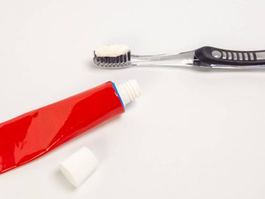 Diş fırçasını diş macunu ve beyaz bir yüzeyde kırmızı bir diş macunu tüpüyle yakından çek. Diş fırçasının siyah tüyleri ve temiz bir sapı var. Kırmızı tüp açık, yanında da kapağı var. Görüntü temiz, parlak bir estetiğe sahip. 