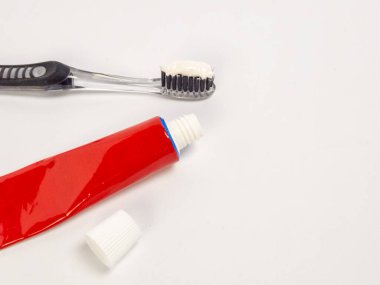 Kapağı açık kırmızı diş macunu tüpü ve diş fırçasıyla yakın çekim bir stüdyo fotoğrafı. Diş fırçası sol alt köşesinde diş macunu tüpü olan üst köşeye yakın bir yerde, hepsi temiz beyaz bir arka fona karşı kurulmuş. Th