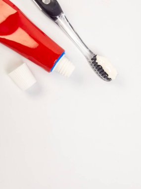 Açık kapaklı kırmızı ve beyaz bir diş macunu tüpünün yakın plan çekimi, ve beyaz diş macunlu temiz bir diş fırçası, düz beyaz bir yüzey üzerine dizilmiş. Görüntü iyi aydınlatılmış, temiz ve minimalist bir estetik. Bu resim wo