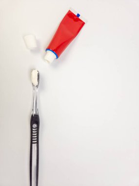 Yakın çekimde diş macunu sürülmüş temiz bir diş fırçası, kapağı çıkarılmış kırmızı bir diş macunu tüpü ve kapağın kendisi görülüyor. Kompozisyon parlak, temiz beyaz bir arkaplan üzerine kurulmuş. Görüntü diş sağlığı için uygundur,