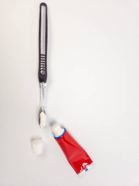 Diş fırçasının tepesinden aşşağıya diş macunu ile kapağı açık kırmızı bir diş macunu tüpünün yanında. Sahne, temiz beyaz bir arka plana göre ayarlanmış. Bu da ağız sağlığı ve diş bakımına odaklandığını gösteriyor. Kompozisyon basit, Em.