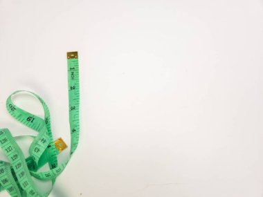 Yeşil bir mezura beyaz bir yüzeyde yatıyor, bant kısmen sökülmüş ve ölçüm işaretlerini gösteriyor. Kaset, çerçeve boyunca çaprazlama yerleştirilerek dinamik bir kompozisyon oluşturuyor. Resmin odak noktası modada kullanılan araçlar. 