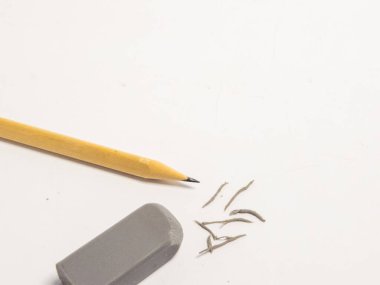 Keskinleştirilmiş uçlu sarı bir kalem, gri bir silgi ve temiz, beyaz bir yüzeye saçılmış küçük bir kalem talaşı içeren yakın plan bir fotoğraf. Resim parlak bir şekilde ışıklandırılmış, nesnelerin dokularını ve detaylarını vurguluyor. Bu natürmort kompozisyon