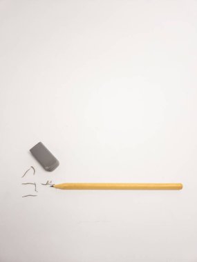 Yakın plan çekimde beyaz bir yüzeyde duran tahta bir kalem ve gri bir silgi bulunur. Kalem kurşun talaşları silginin yanına saçılmış. Silme ve düzeltme olduğunu gösteriyor. Basit kompozisyon ve nötr arkaplan temiz, minimalist bir aesth oluşturur.