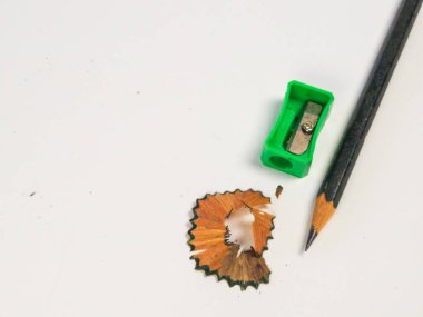 Keskinleştirilmiş bir kalem, yeşil bir kalem açacağı ve beyaz arka planda ahşap talaşı. Görüntü, siyah kalem, yeşil bileyici ve ahşap talaşlarının sıcak tonları arasındaki kontrastı vurgular. Kompozisyon si