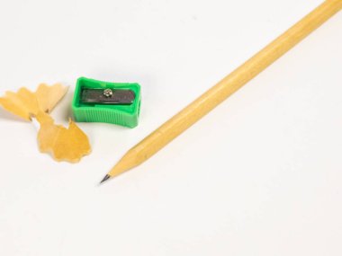 Keskinleştirilmiş bir kalem, yeşil bir kalem açacağı ve sade beyaz bir arka planda ahşap talaşı. Görüntü, kalemin keskin noktasını, talaş dokusunu ve keskinleştiricinin detaylarını gösteriyor. Kompozisyon basit ve temiz, vurgulu.