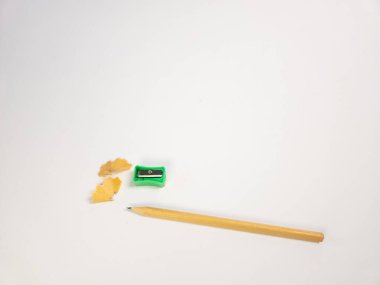 Bu resimde bir kalem, bir yeşil kalem açacağı ve el değmemiş beyaz arka planda ahşap talaşları görülüyor. Kompozisyon basit ve minimalist, kırtasiye malzemelerini vurguluyor. Görüntü iyi aydınlatılmış ve odak noktası keskin.